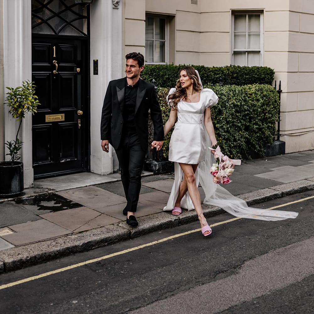 Groom in a black shirt, suit and loafers crossing the road with his bride in a short wedding dress with puff shoulder detail 