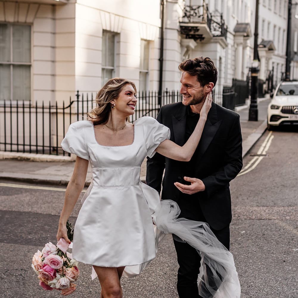 Bride in a short wedding dress wearing pink mules and holding a blush, peeled back rose bouquet embracing her grooms face 