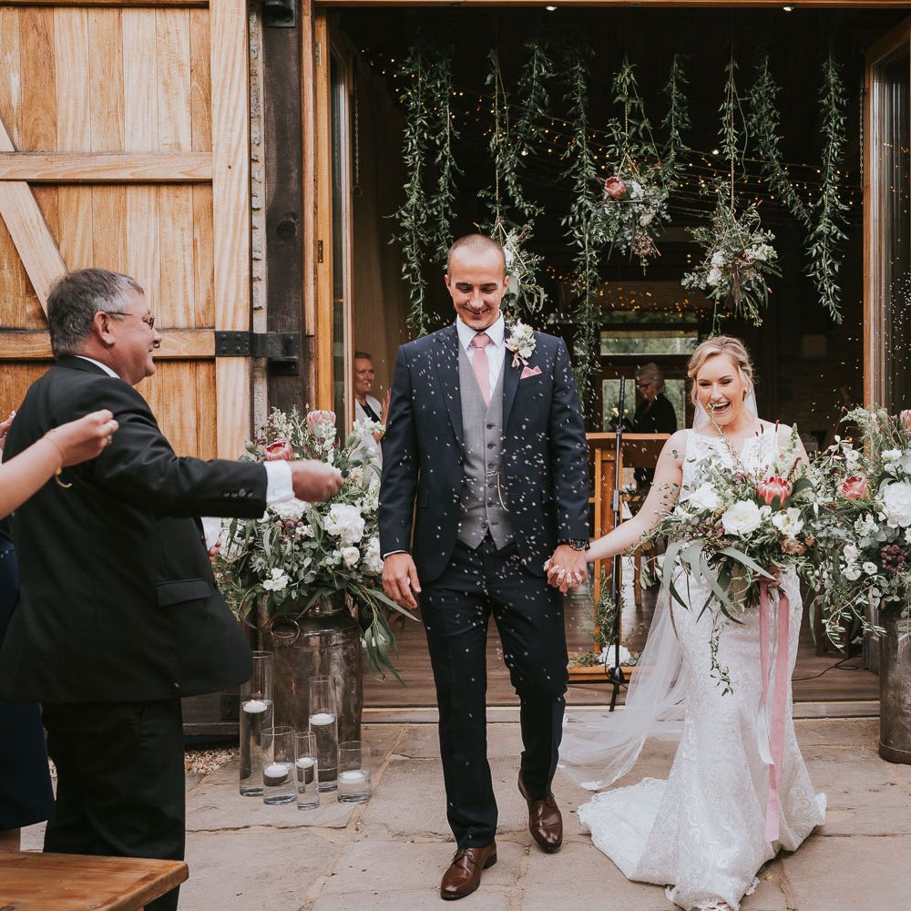 Groom in dark three piece suit with grey waistcoat and pink tie holding hands with bride in lace Justin Alexander wedding dress holding pink and white bridal bouquet walk back down the aisle whilst guests throw confetti at Tythe Barn wedding 