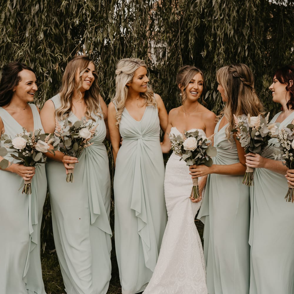 Bride stands with her bridesmaids who wear sage green dresses and carry white floral bouquets | Mark Bamforth Photography
