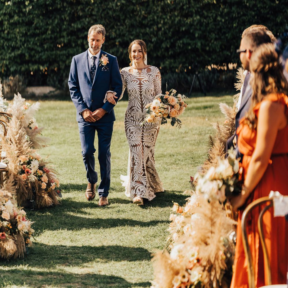 Bride in lace wedding dress walking in outdoor setting with father navy three piece suit 