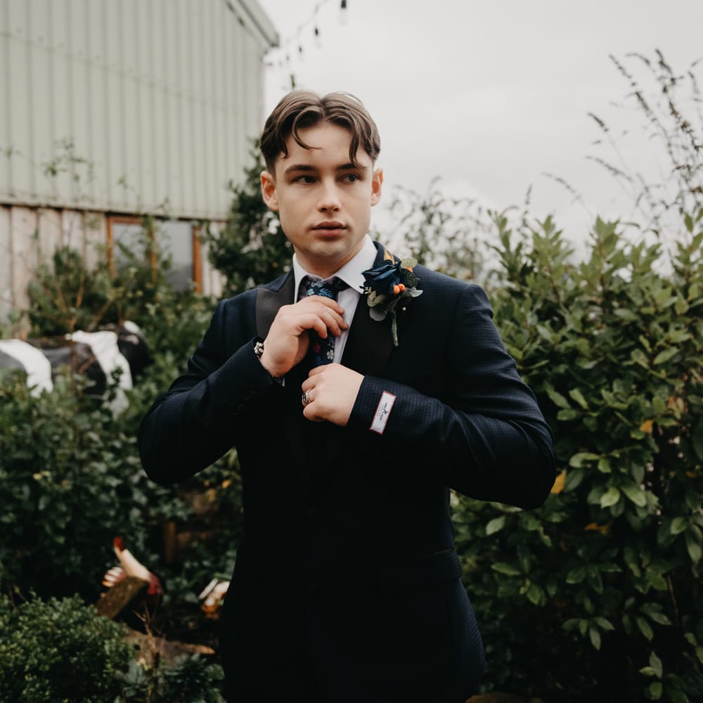 Groom in dark blue suit, floral tie and floral buttonhole outside for rustic farm wedding