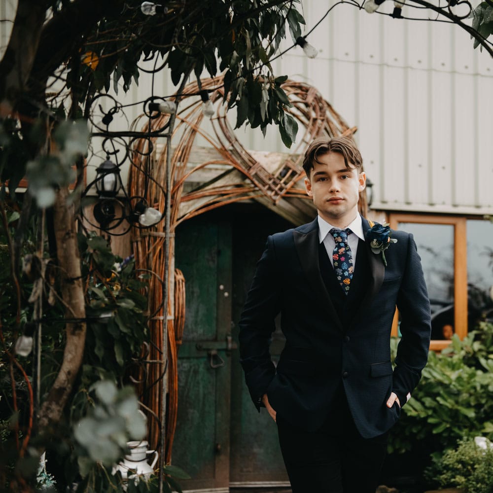 Groom in blue suit, floral tie and rose buttonhole walks through garden for rustic farm wedding in Lancashire