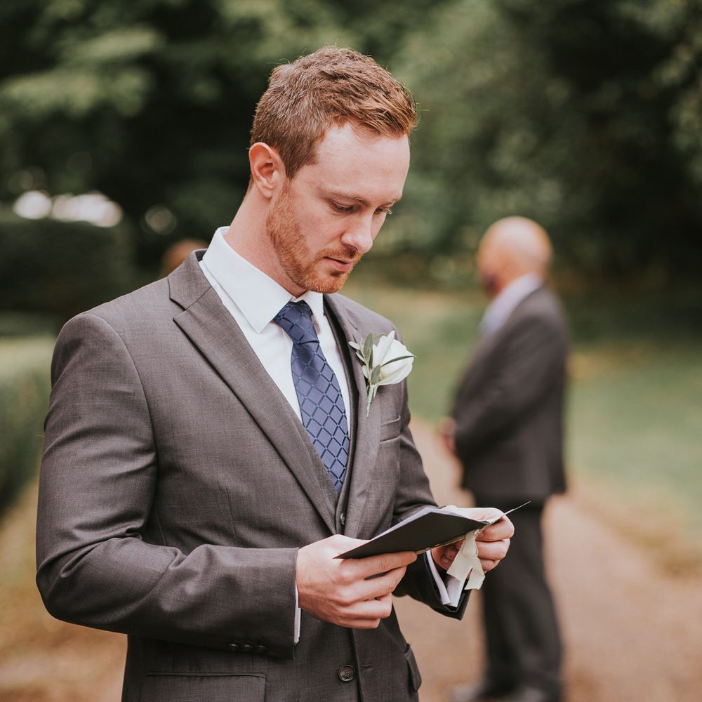 Groom in grey three piece suit, blue tie and whit rose buttonhole reads vows before outdoor wedding ceremony