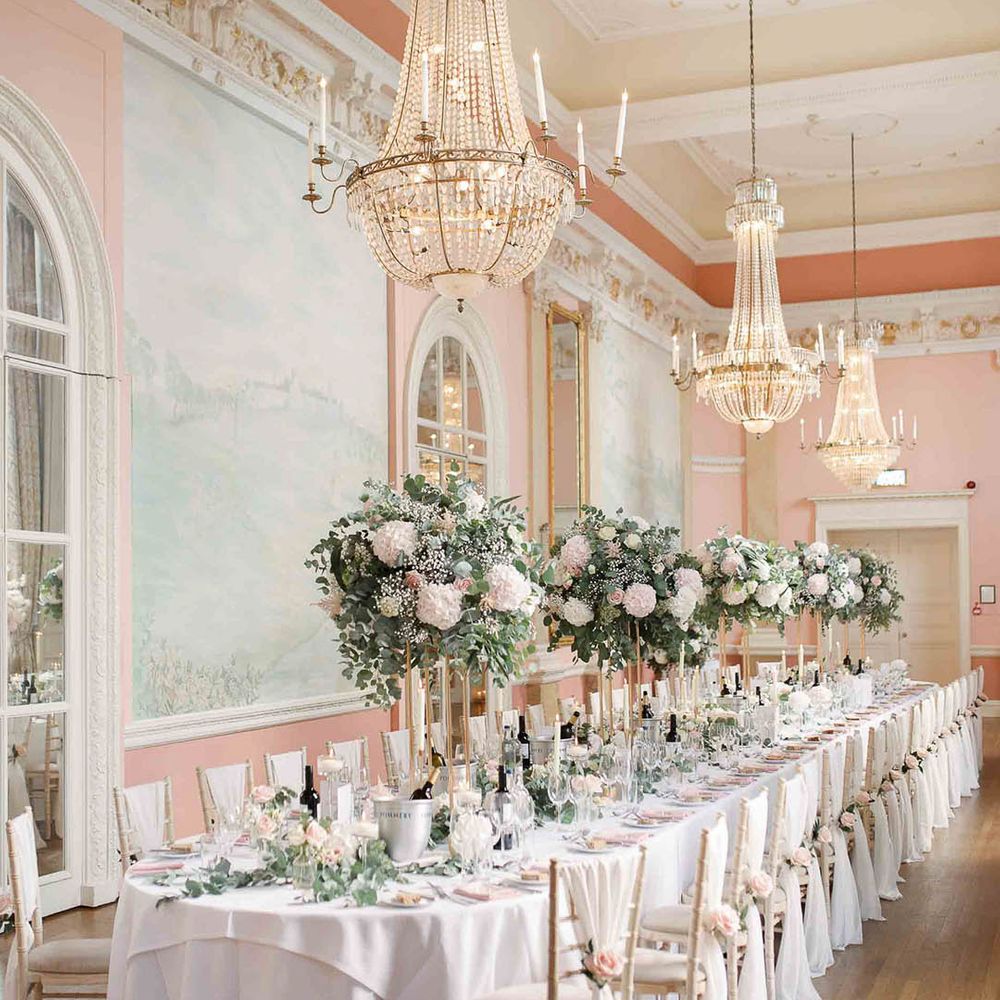 Danesfield House reception complete with grand chandeliers, long table lined with white cloth, large floral bouquets filled with blush pink and white flowers