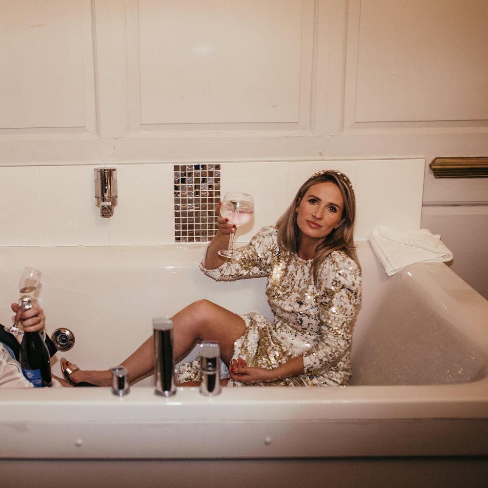 Bride & groom sit in bathtub on their wedding day and hold champagne glasses 