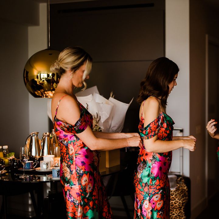 Bridesmaids in bright floral dresses zip each other up as they get ready for the wedding day