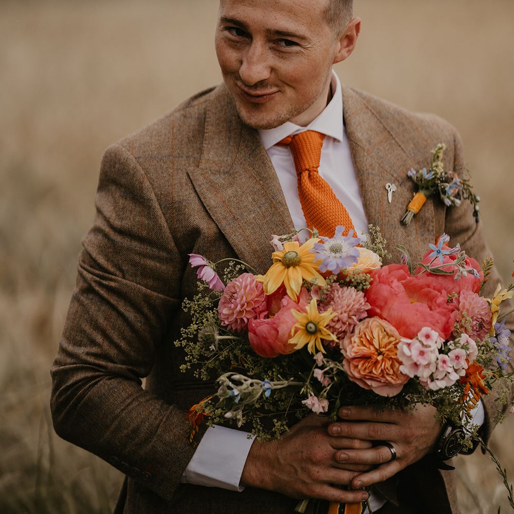 Groom in brown suit and orange tie makes a cheeky face as he holds the bridal bouquet 