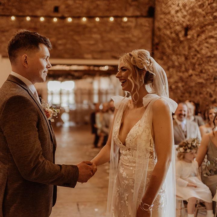 Bride in Elsie Made With Love wedding dress holding hands with groom at the altar with groom in brown three piece suit 