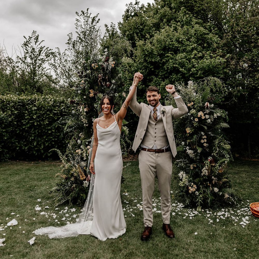 Bride in a satin wedding dress with pearl veil holding hands with her groom in a stone suit at their outdoor wedding ceremony with greenery column flower arrangements 