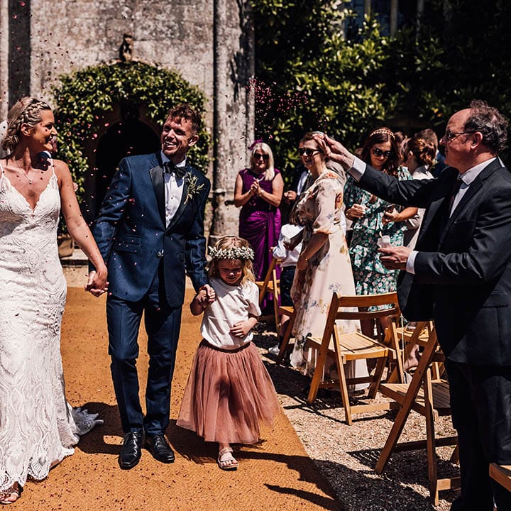 Bride in lace wedding dress and groom in blue tuxedo with their daughter in a pink tulle skirt and white flower crown have confetti exit from their wedding 