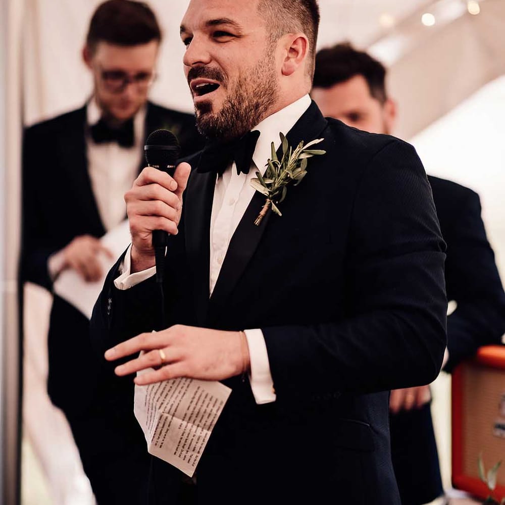 Best man in black tie reads out his speech wearing simple green foliage buttonhole 
