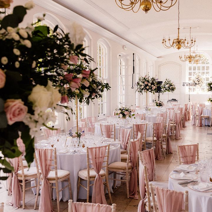 White reception room at Goldney Hall with chair backs decorated with pink chiffon material and tables decorated with tall pink rose centrepieces and gold chandeliers 