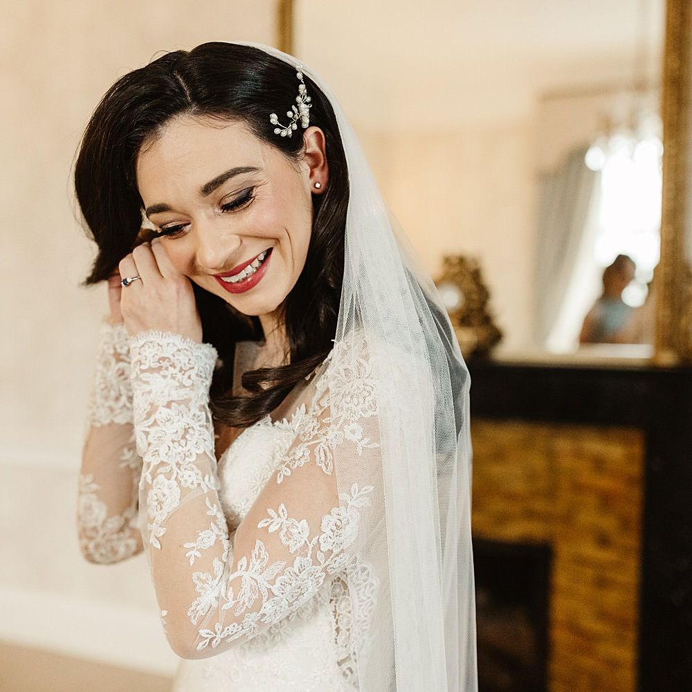Bride puts in earrings as she wears long sleeved lace gown complete with her dark hair in loose curls 