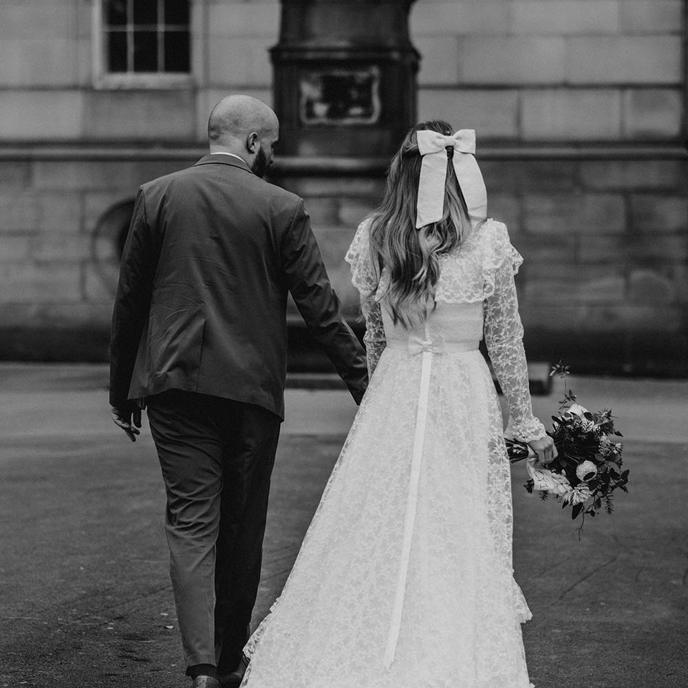 Bride wears bow in her long hair as she walks hand in hand with her groom in black and white image