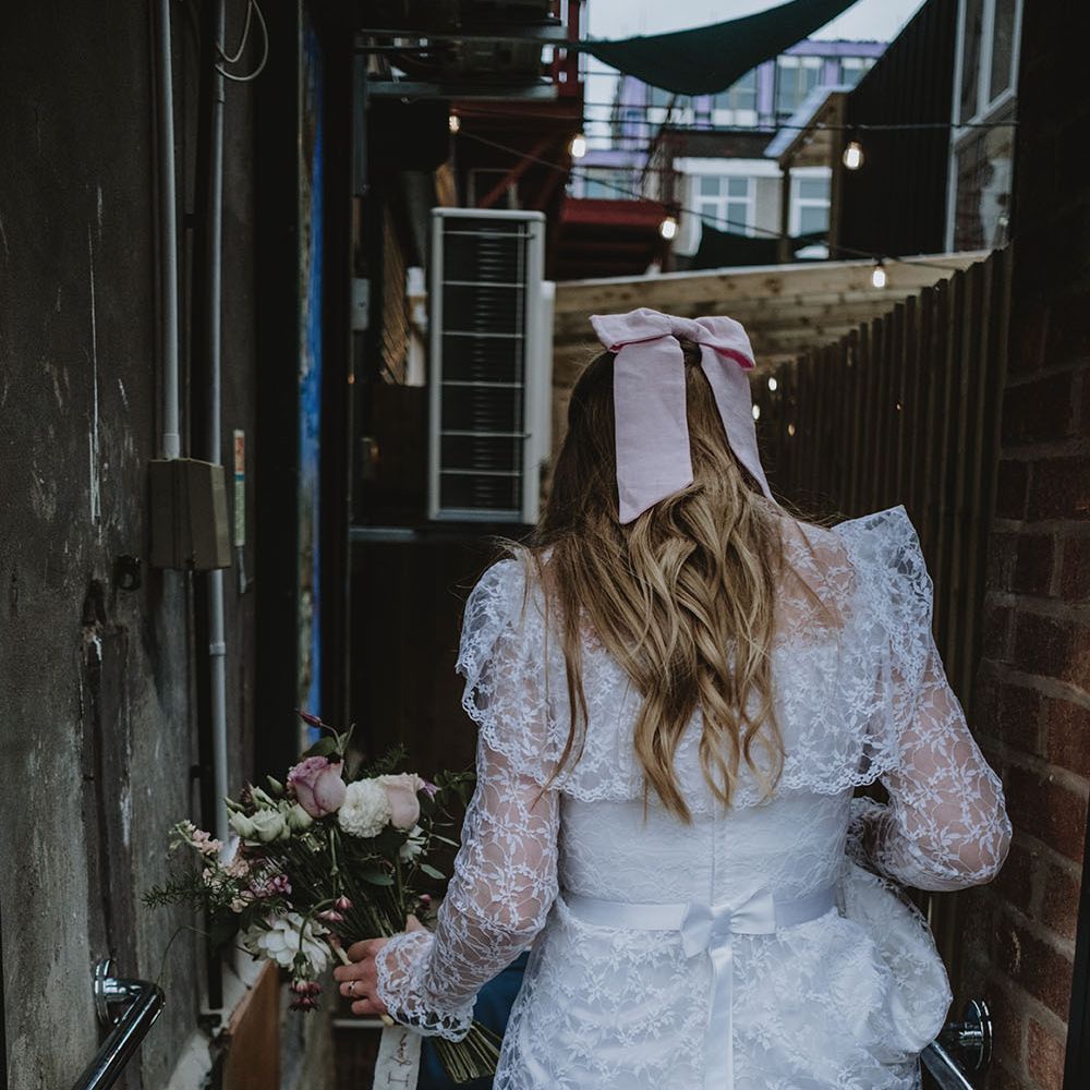 Bride wears lace vintage wedding dress from Oxfam with pink bow in her long blonde curled hair whilst carrying pink and white floral bouquet