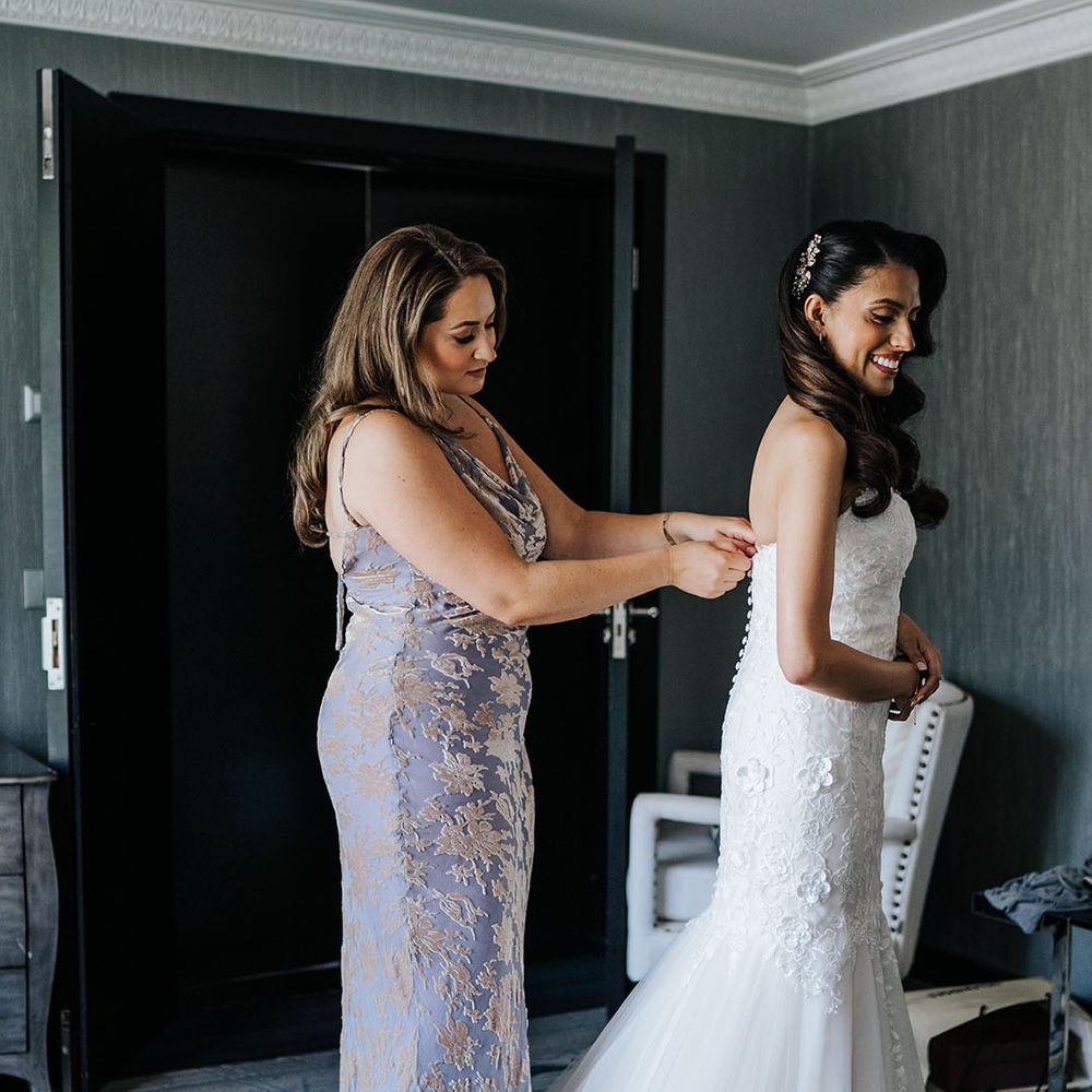 Bridesmaid wears lavender bridesmaid dress and does up buttons to the back of brides lace wedding dress 