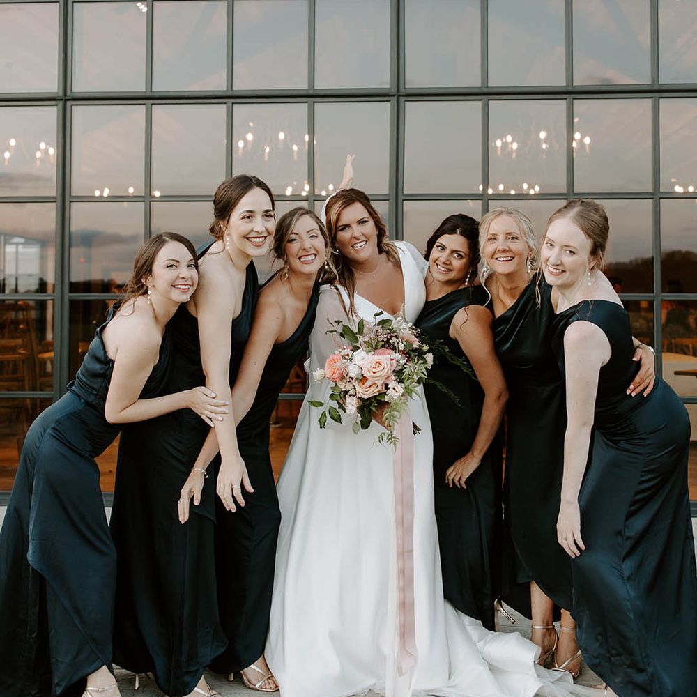 Bride holds pastel bouquet tied with pink ribbon and stands with her bridesmaids in green satin bridesmaid dresses with one shoulder