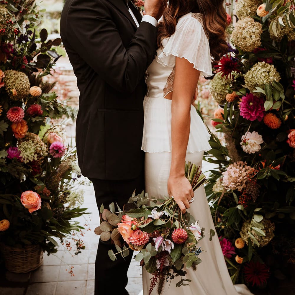 Bride and groom embracing by large luxe floral arch with white and pink carnations, garden roses, red strawflower, eucalyptus, baby’s breath and various dried flowers 