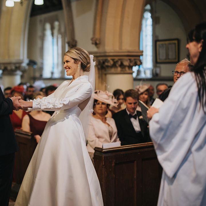 The groom in black tie smiles facing the bird holding both of her hands as she laughs wearing a long sleeve with ruffle detail wedding dress