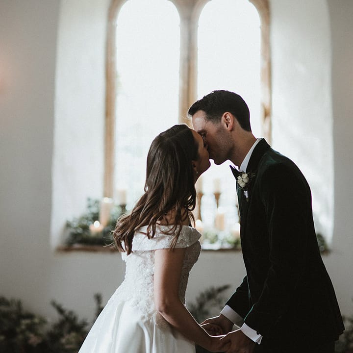 Bride in lace off the shoulder wedding dress from Pronovias wedding dress sharing a first kiss with the groom in a velvet green tuxedo 