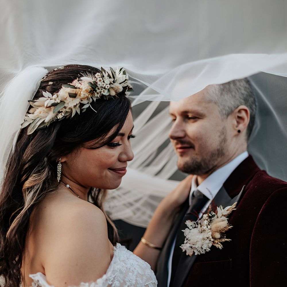 The bride wears an elegant white boho flower crown and embraces the groom in a velvet suit jacket with dried flower buttonhole under the bride's veil