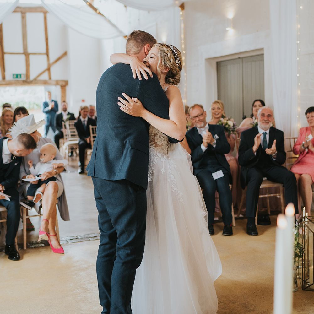 Bride with blonde braided hairstyle with sparkly headpiece embraces the groom in a navy suit at the altar in front of the wedding guests 
