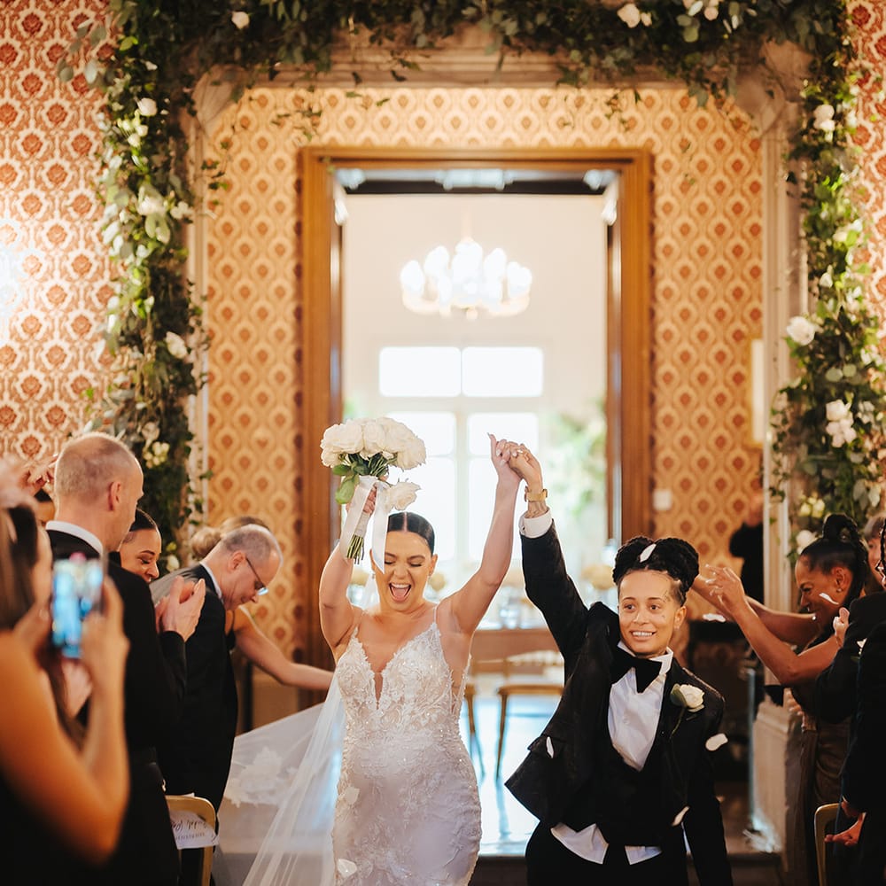 Bride in black snakeskin print tuxedo with black bowtie and white rose boutonniere and bride in lace sleeveless v neck Enzoani wedding dress with tulle finish and church length veil holding white garden rose bridal bouquet doing confetti exit at black tie lesbian wedding at Grittleton House