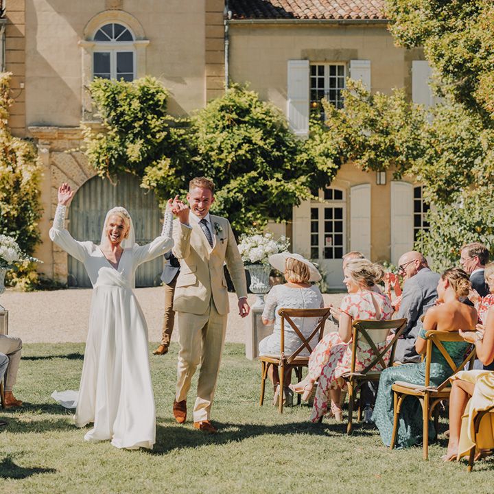 Bride in an Emma Beaumont wedding dress and groom in a beige suit descending down the aisle at Château de Malliac