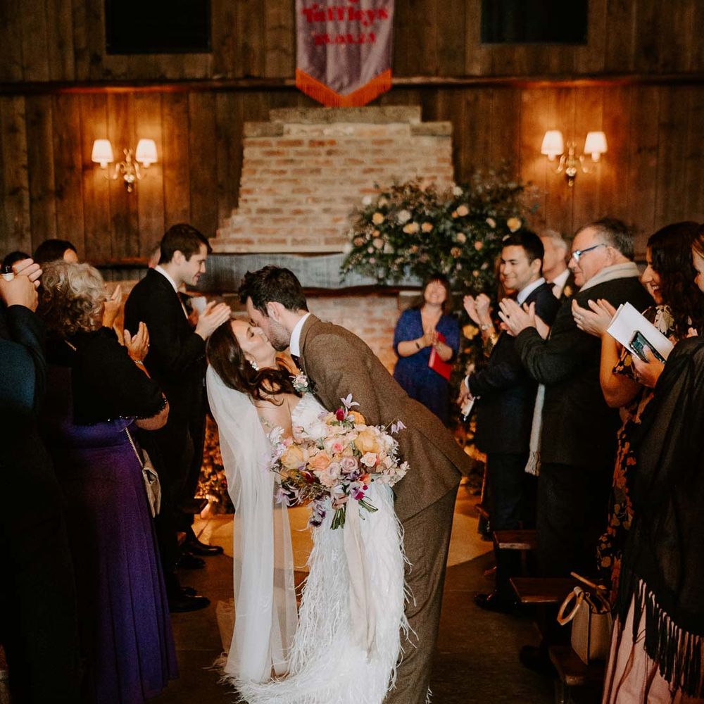 Groom in chocolate brown grooms suit kissing bride in strapless ivory fringed crepe gown and chapel length veil whilst walking down the aisle at Willow Marsh Farm Loughborough 