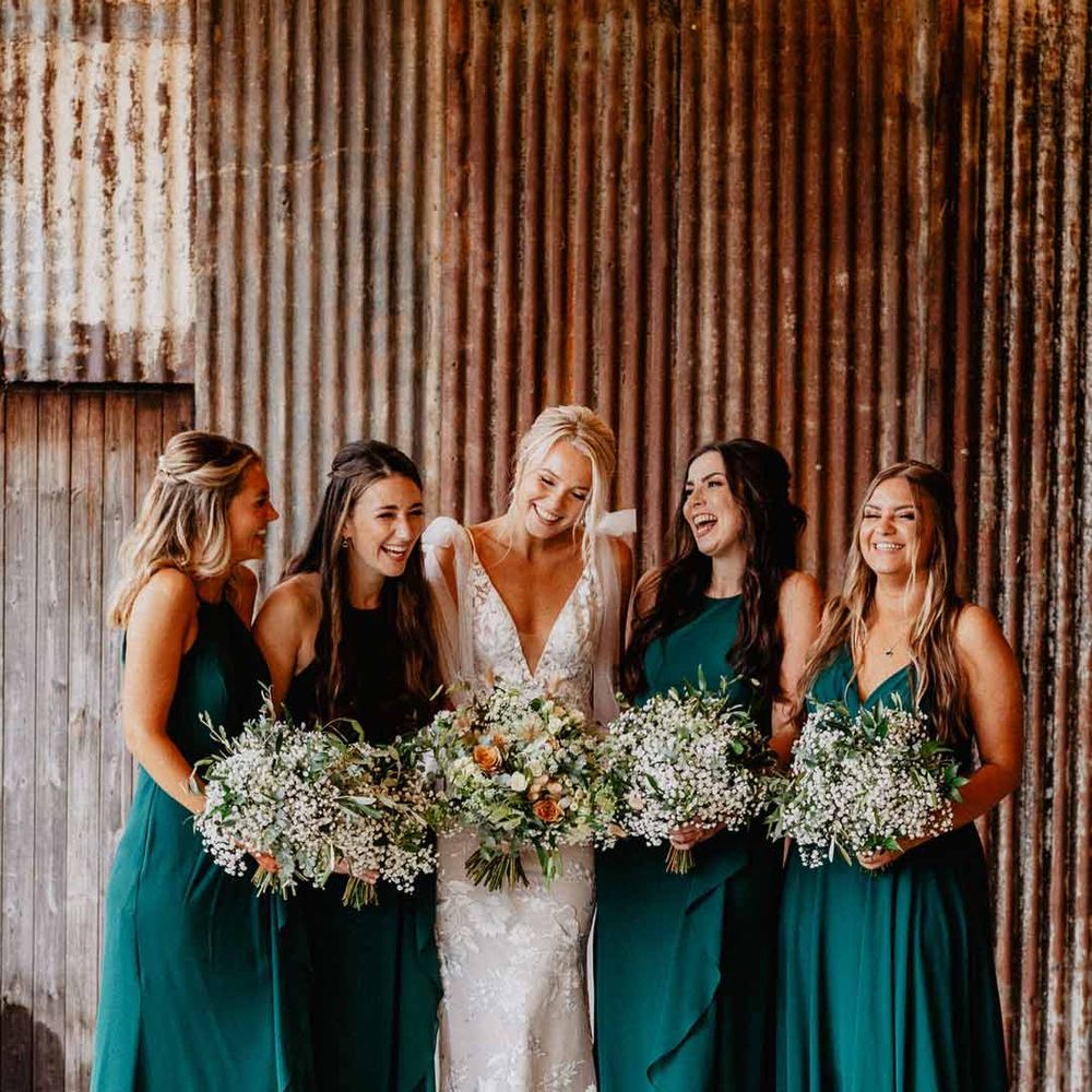 Bride in low v neck lace wedding dress standing with bridesmaids in bottle green mismatched bridesmaid dresses