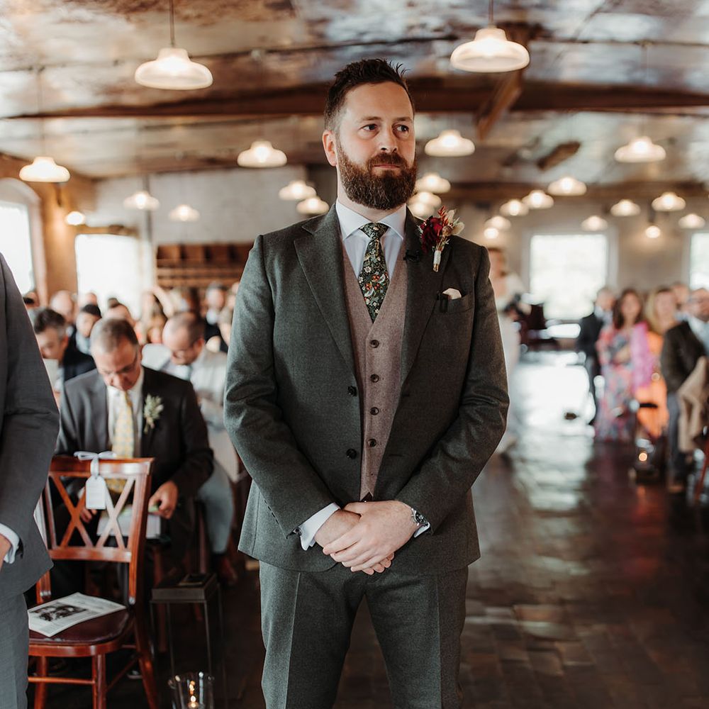 Groom in green wedding suit with grey waistcoat and floral patterned tie waiting for the bride at the end of the aisle 