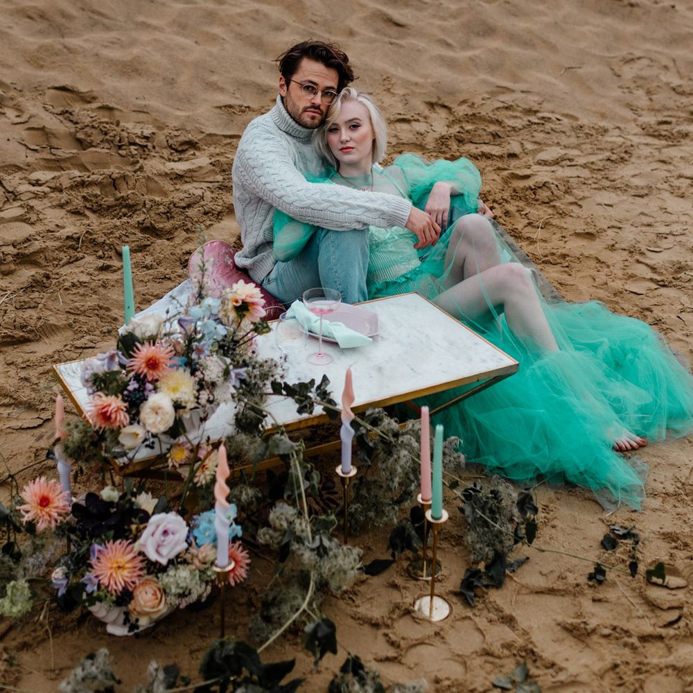 Bride in a pastel green wedding dress sitting on the sand by her picnic table decorated with colourful flowers and candles with her groom in a grey jumper 
