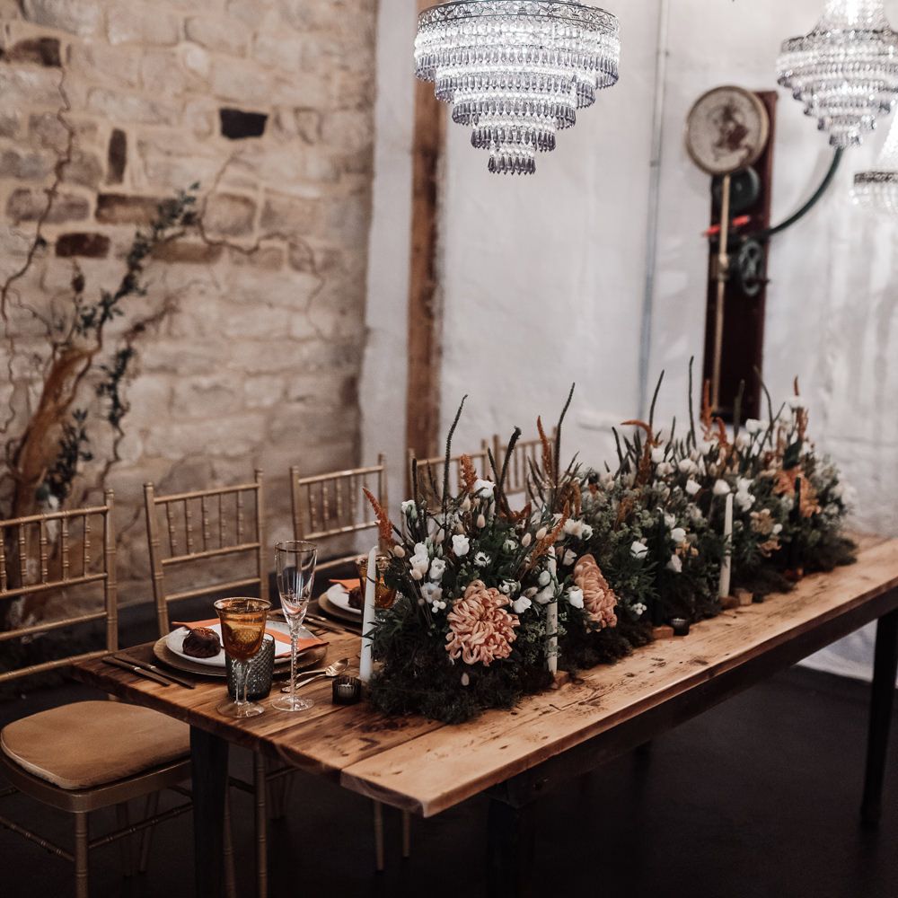 Chandelier light installation over a wooden table with orange flower and foliage table runner of flowers at Ponden Mill wedding venue 