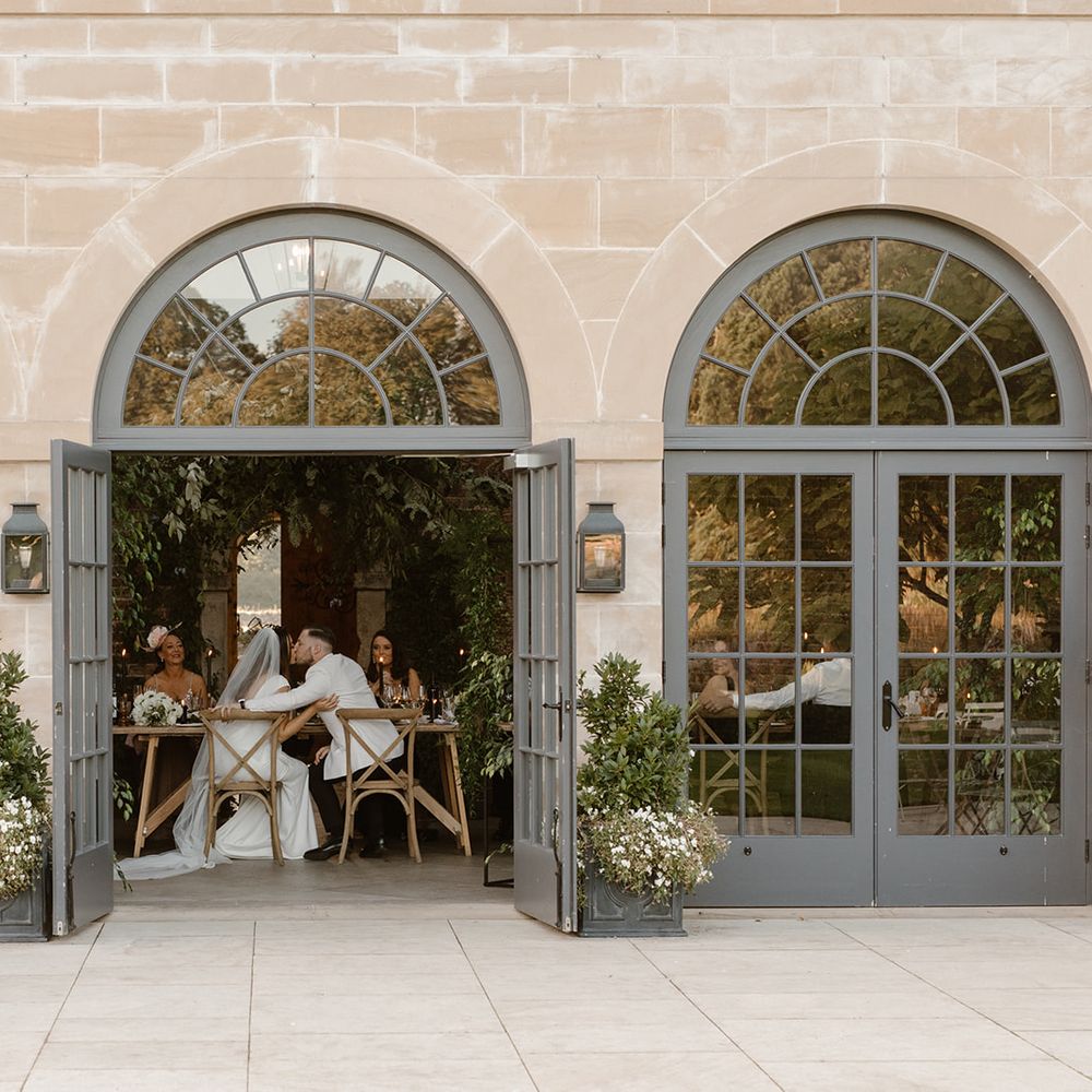 Bride and groom embrace sat at wedding table through open doors in the Fig House at Middleton Lodge