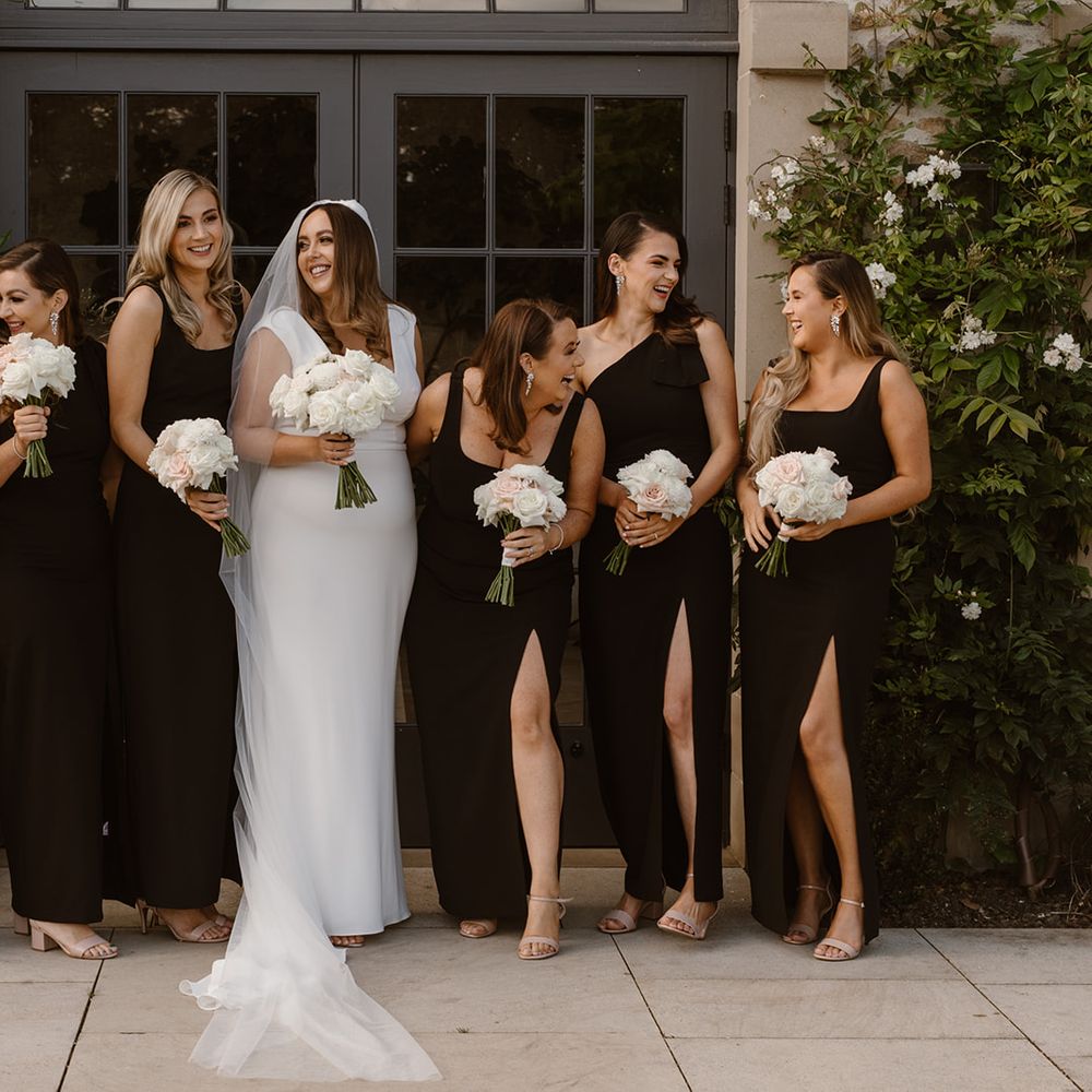 Bride in white Made With Love wedding dress poses outside with bridesmaids in long black bridesmaid dresses all holding white and blush pink rose bouquets