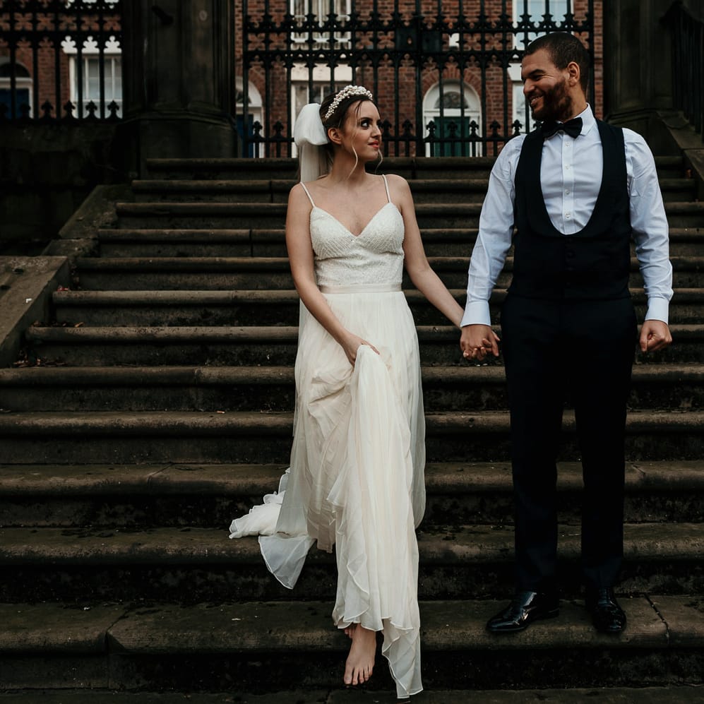 Groom in a horseshoe waistcoat and bow tie walking down the steps holding hands with his bride in a thin strap wedding dress and bow veil 