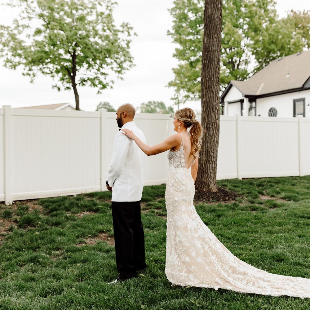 Bride with ponytail in embroidered Made With Love wedding dress with train taps groom in white tuxedo jacket on the shoulder for first look at backyard wedding