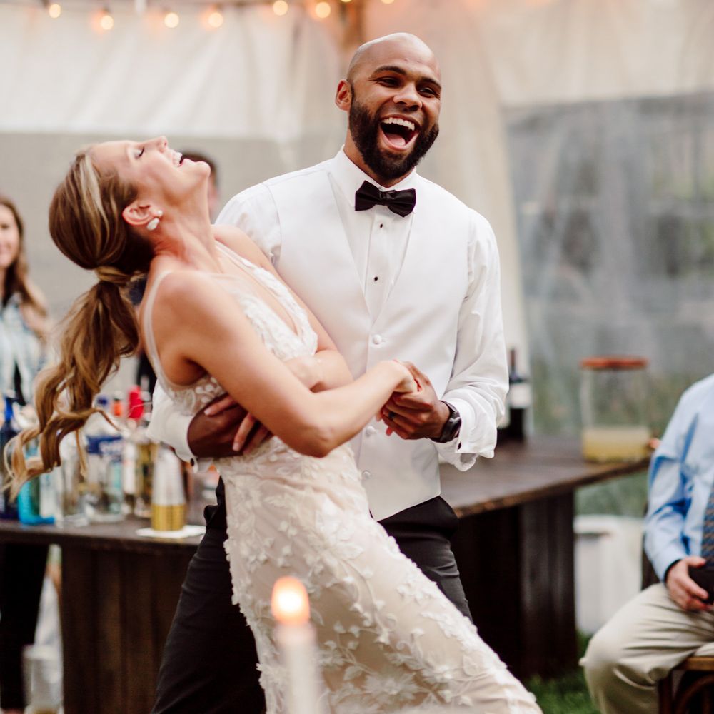 Laughing groom in white waistcoat, white shirt and black bow tie dances with laughing bride in white Made With Love wedding dress in fairy lit marquee during backyard wedding