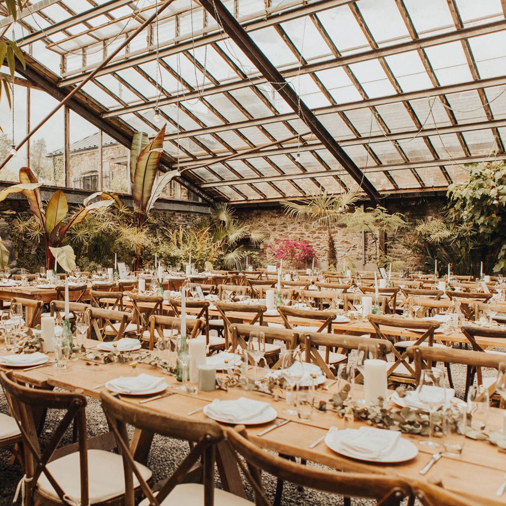Interior of glasshouse at Anran Devon for wedding breakfast with long wooden tables, wooden chairs, candles and tableware, and large fern and palm plants