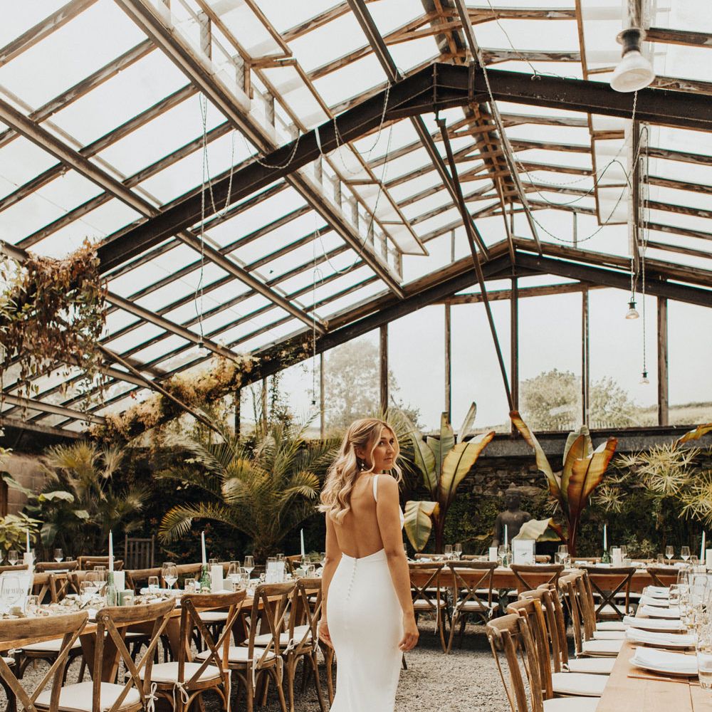 Bride in white open back Made With Love wedding dress stands in glasshouse at Anran Devon for wedding breakfast with long wooden tables, wooden chairs, candles and large fern plants 