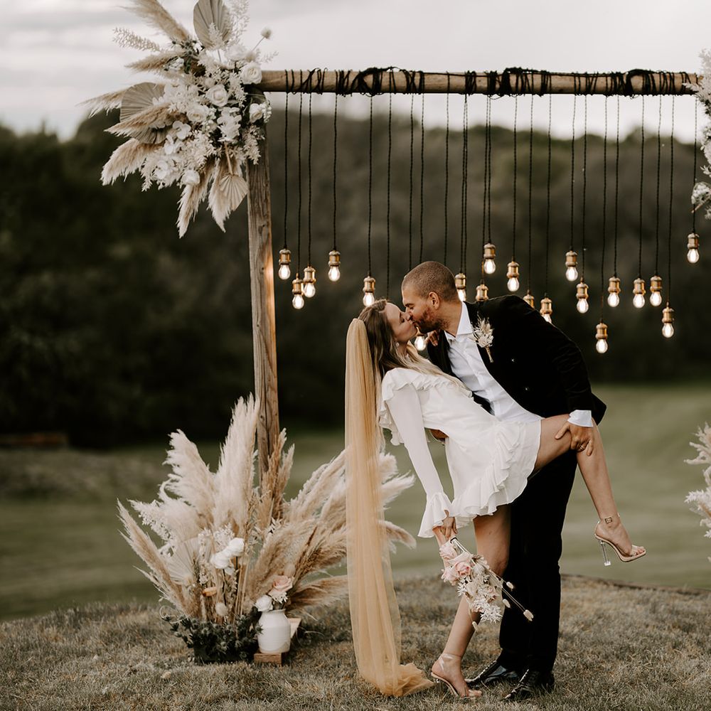 Groom in a black suit leaning his bride over in a short wedding dress and long coloured veil in front of a wooden altar decorated with vintage bulbs and dried flowers 