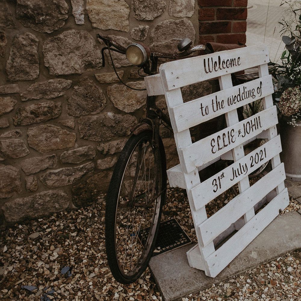 White wooden pallet wedding welcome sign leaning on bike next to milk churn full of florals for Isle of Wight wedding with macrame wedding decor