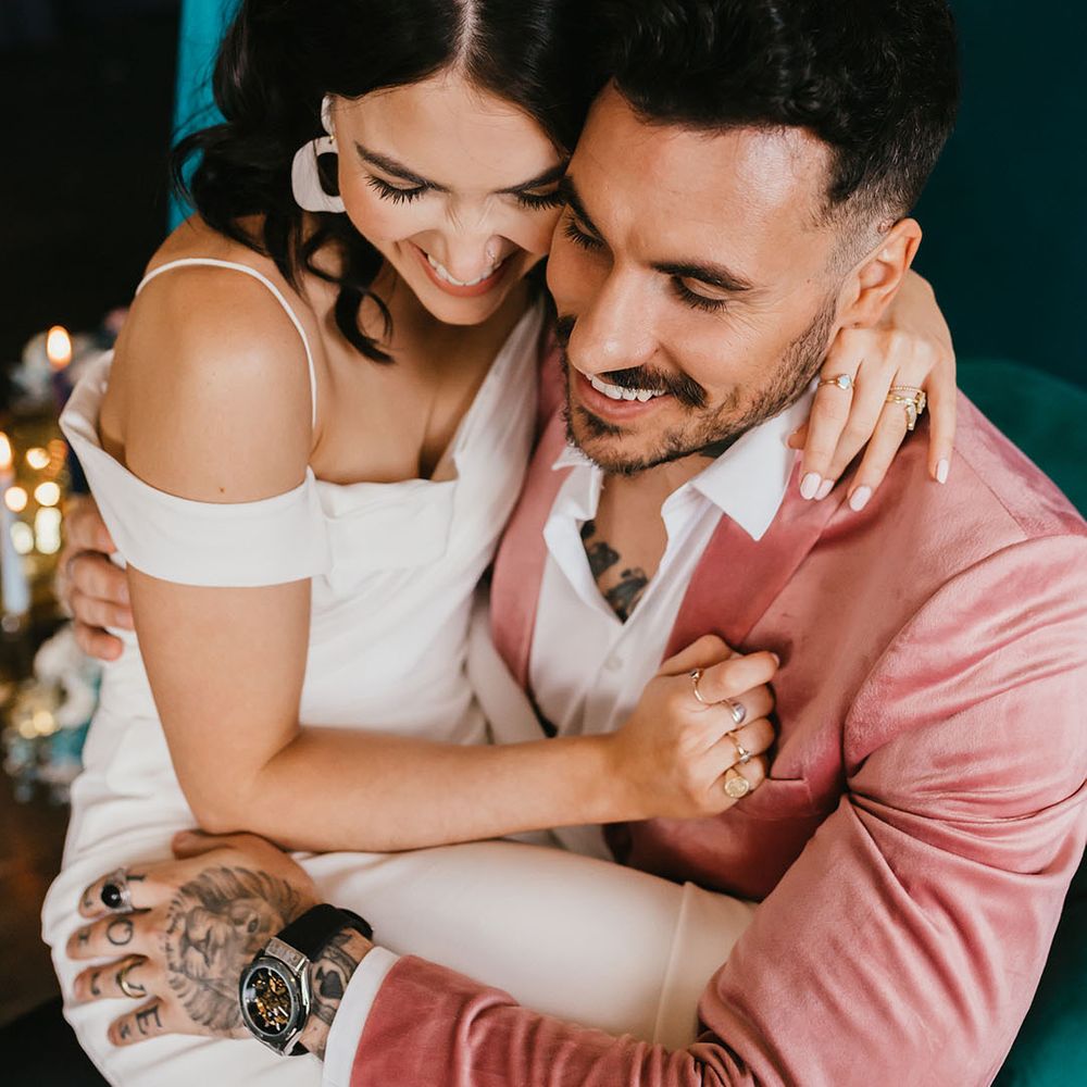 Bride with white nail polish and ring stacks embracing her tattooed groom in a pink velvet jacket 