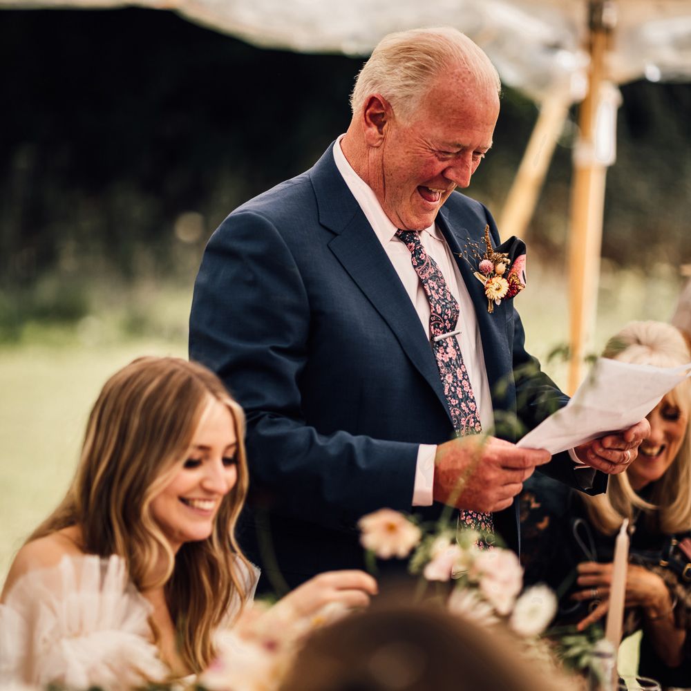 Father of the bride in blue suit and patterned ties makes speech during marquee wedding reception in Cornwall
