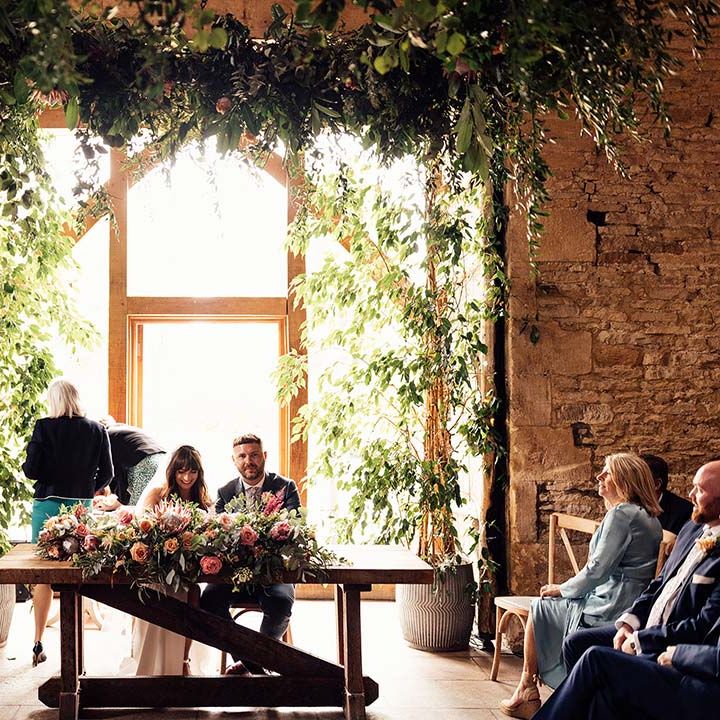 Bride & groom sit in front of large barn window during wedding ceremony as they sign their marriage certificate 