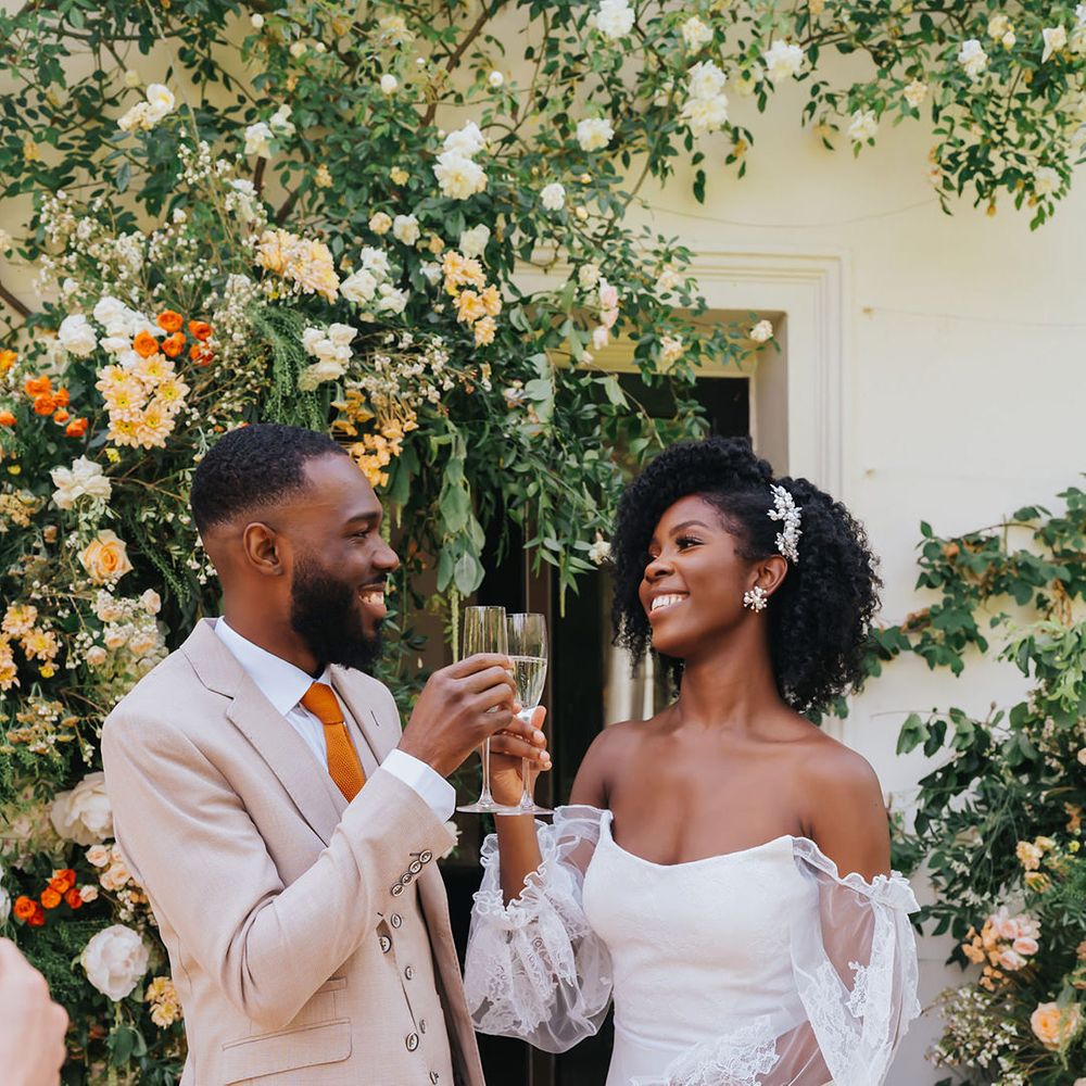 Black bride and groom in a beige suit and strapless wedding dress with sheer sleeves toasting champagne at their outdoor wedding reception 