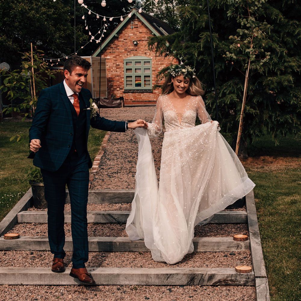 Bride in long sleeve v neck wedding dress and flower crown runs down garden stairs holding groom in blue checked three piece suit at garden wedding reception