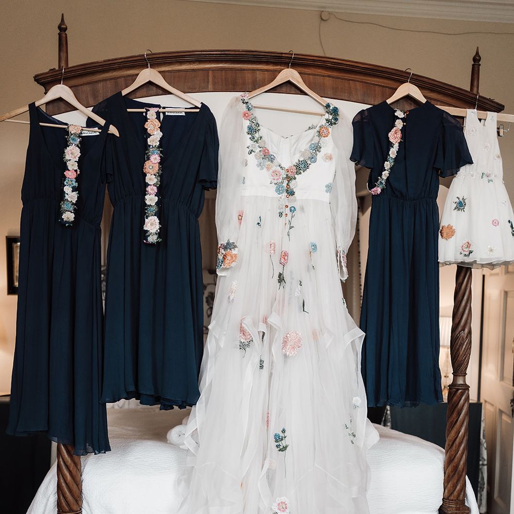 Long sleeve Victoria Sanders wedding dress with floral embroidery and train hung on four poster bed with navy bridesmaid dresses