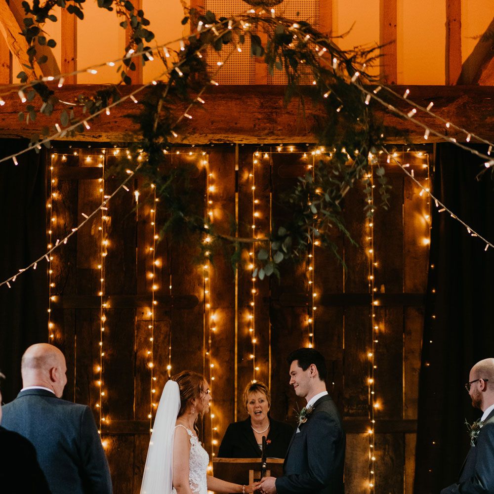 Bride in lace and tulle wedding dress and veil stands in front of fairy light wall with groom in dark suit for rustic wedding ceremony